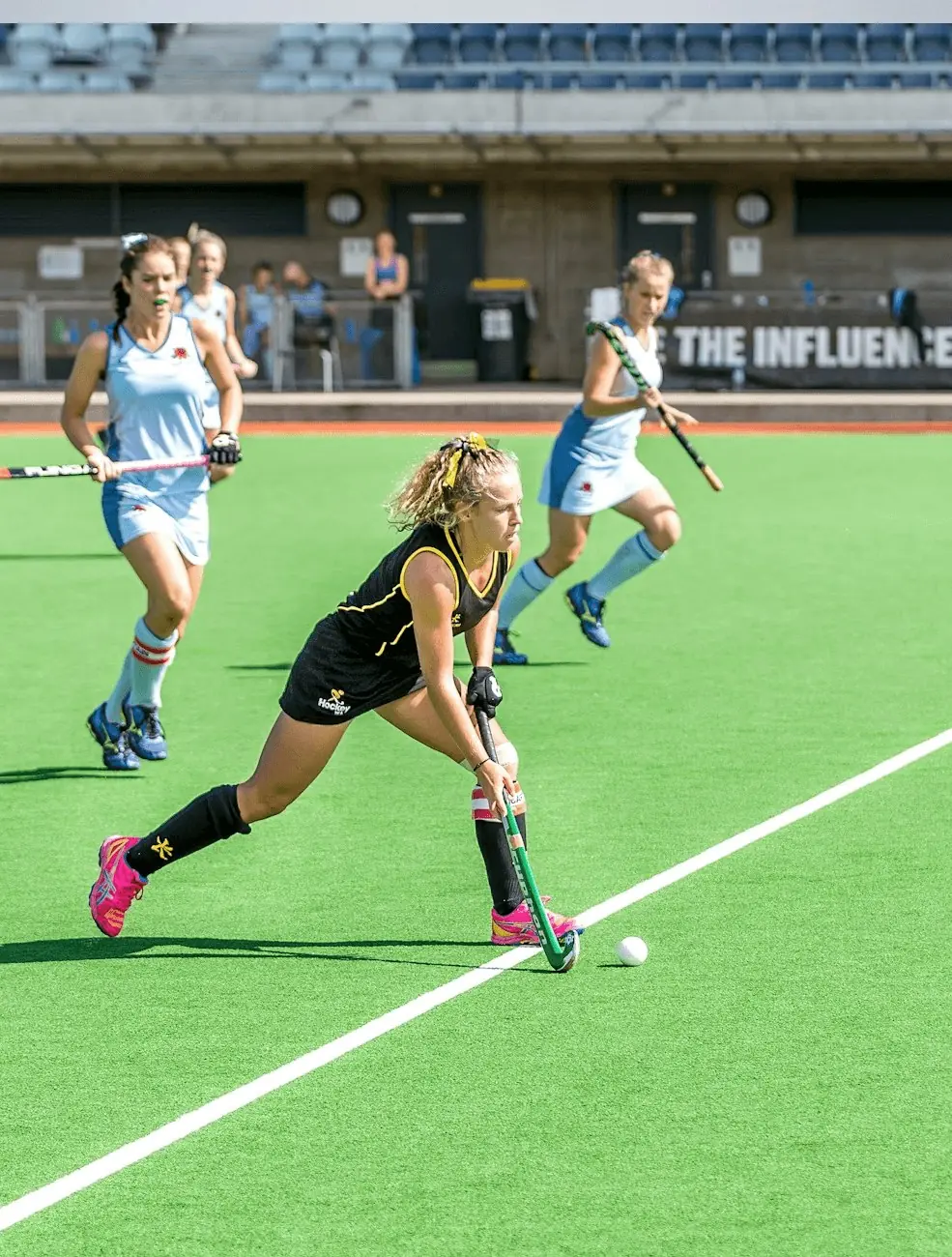 Hockey sur gazon sur un terrain vert : une joueuse en noir avance avec la balle.