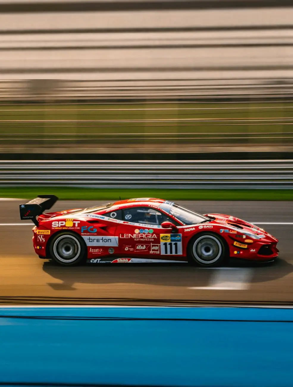 A red GT race car at speed on a circuit, its bodywork covered in sponsor logos.